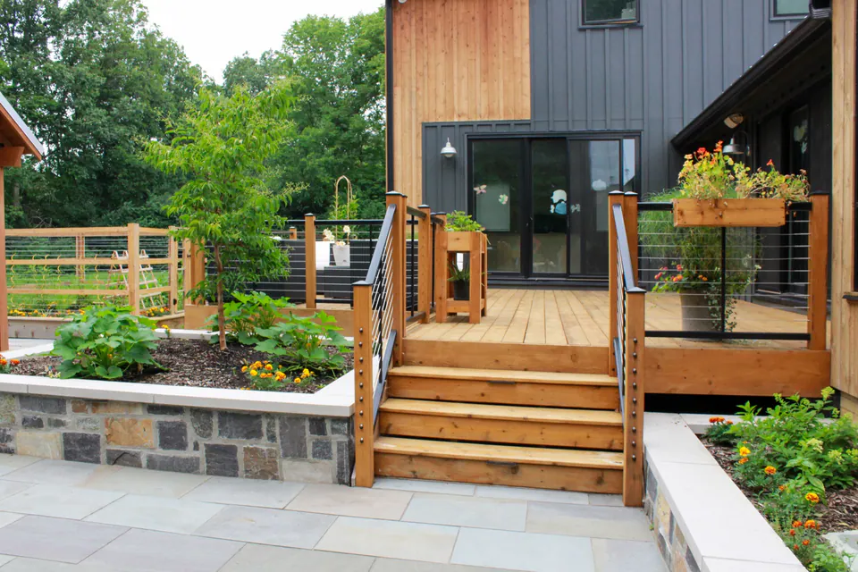 Deck stairs and stone patio with raised beds, house siding, railings, planter box, and garden planting.